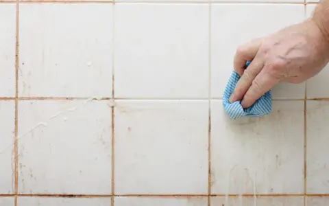 Hand reinigt schmutzige weiße Wandfliesen mit einem blauen Tuch.