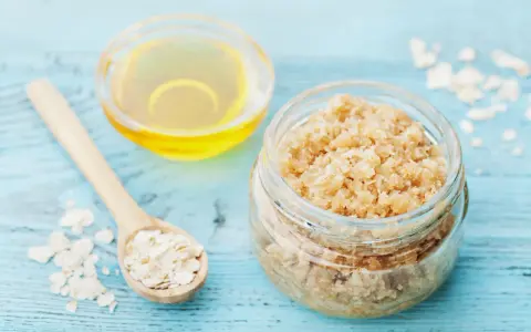 Zutaten für ein **Gesichtspeeling selber machen**: Haferflocken auf einem Holzlöffel, ein Glas mit Peeling und eine Schale mit Öl.