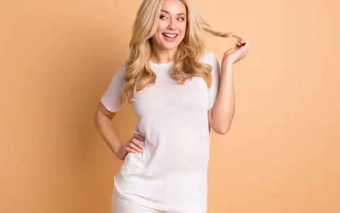 Eine lächelnde Vanilla Girl mit blonden Locken spielt mit einer Haarsträhne. Sie trägt ein weißes T-Shirt und weiße Hose.