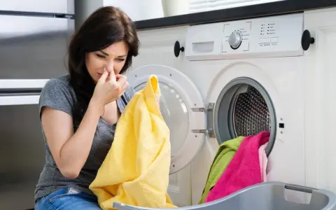 Frau hält sich die Nase zu, weil die Waschmaschine stinkt. Gelbes Handtuch in der Hand, blickt sie angewidert auf die Wäsche.