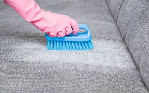 Hand in pink glove uses blue brush to scrub grey fabric, showing how to remove blutflecken.