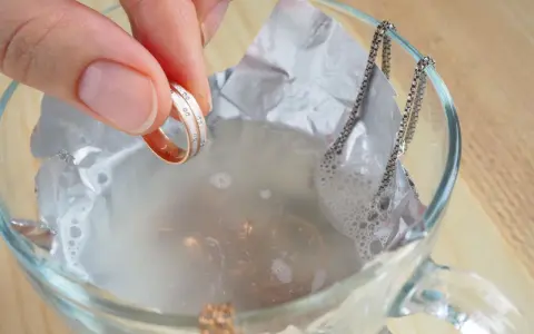 Schmuckreinigung mit Hausmitteln: Eine Hand hält einen Ring, während eine Silberkette in einer Schüssel mit Wasser und Alufolie liegt.