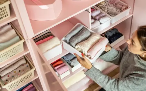 Frau sortiert Kleidung in einem rosa Kleiderschrank. Sie zeigt, wie man den Kleiderschrank organisiert, mit gefalteten Pullovern in weißen Boxen.