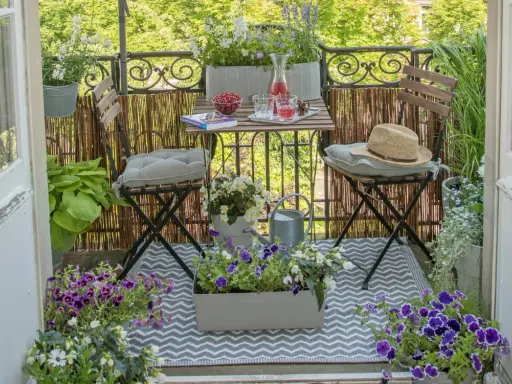 Gemütliche Balkon Ideen: Ein kleiner Tisch mit Getränken und Kirschen, zwei Stühle mit Kissen, Blumen und eine Gießkanne laden zum Verweilen ein.