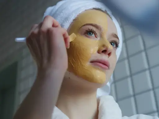 Junge Frau mit Handtuch auf dem Kopf trägt eine Kurkuma-Maske auf. Die Kurkuma Wirkung auf die Haut ist bekannt für ihre entzündungshemmenden Eigenschaften.