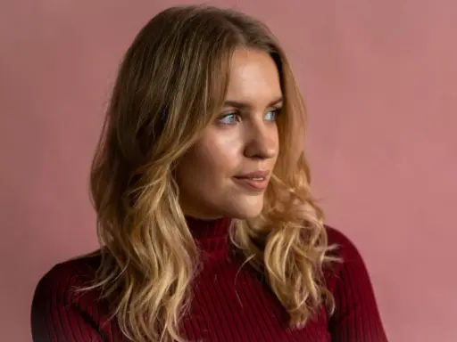Blonde Frau mit einem **kitty cut**-Haarschnitt, trägt einen roten Rollkragenpullover vor rosafarbenem Hintergrund.