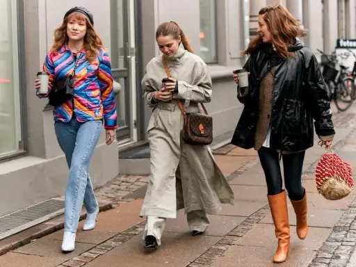 Drei Frauen spazieren mit Kaffee. Die mittlere trägt weite Hosen zu Pumps, die rechte Leggings zu Stiefeln. Die linke zeigt, welche Hose zu Boots Damen passt: Jeans zu weißen Stiefeletten.