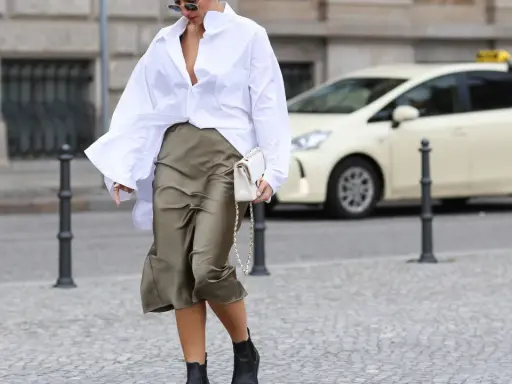 Eine Frau stylt eine weiße Oversize Bluse mit einem Satinrock und schwarzen Stiefeln. Sie trägt eine weiße Tasche.