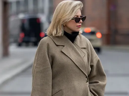 Eine Frau mit blondem Scandi Bob und Sonnenbrille trägt einen beigen Wollmantel und schwarzen Rollkragenpullover.