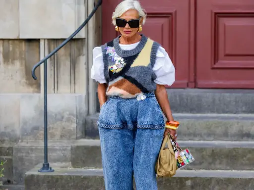 Modische Frau mit weißem Haar, Sonnenbrille und auffälligem Pullunder, der lässig zu Jeans kombiniert wird.