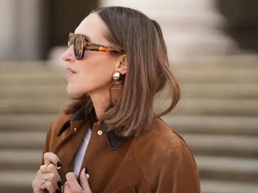 Eine Frau mit braunem Bob und auffälliger Sonnenbrille trägt eine braune Wildlederjacke. Sie erinnert an den Stil von Rodeo Bob.