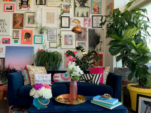 Ein Wohnzimmer im maximalistischen Stil mit einer blauen Couch, vielen Kissen, Pflanzen und einer Wand voller Kunstwerke und Drucke.