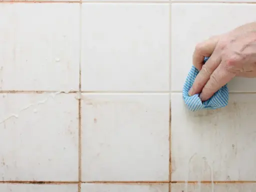 Hand reinigt schmutzige weiße Wandfliesen mit einem blauen Tuch.
