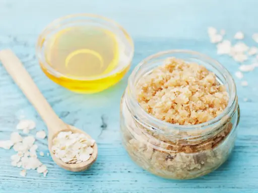 Zutaten für ein **Gesichtspeeling selber machen**: Haferflocken auf einem Holzlöffel, ein Glas mit Peeling und eine Schale mit Öl.
