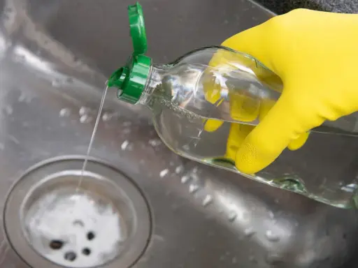 Gelbe Handschuhe halten eine Flasche mit klarem Flüssigkeit, die in ein Spülbecken gegossen wird. Ein Hausmittel Rohrreiniger, der Abfluss wird gereinigt.