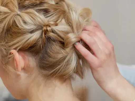 Eine Hand stylt blonde Haare zu verspielten hochsteckfrisuren mittellang. Kleine Zöpfe und gedrehte Strähnen ergeben einen lässigen Look.