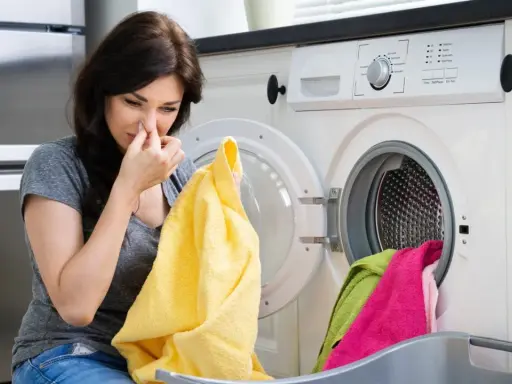 Frau hält sich die Nase zu, weil die Waschmaschine stinkt. Gelbes Handtuch in der Hand, blickt sie angewidert auf die Wäsche.
