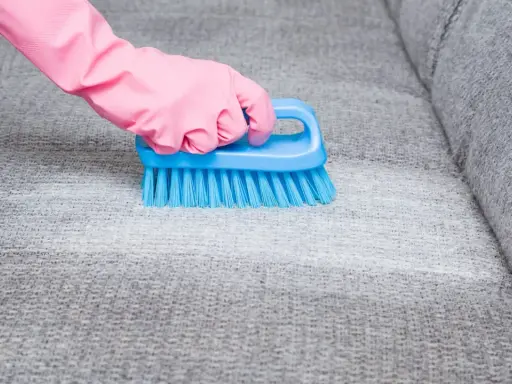 Hand in pink glove uses blue brush to scrub grey fabric, showing how to remove blutflecken.