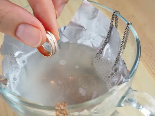 Schmuckreinigung mit Hausmitteln: Eine Hand hält einen Ring, während eine Silberkette in einer Schüssel mit Wasser und Alufolie liegt.