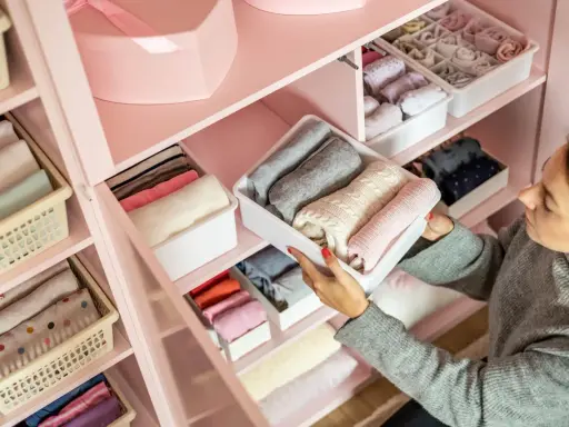 Frau sortiert Kleidung in einem rosa Kleiderschrank. Sie zeigt, wie man den Kleiderschrank organisiert, mit gefalteten Pullovern in weißen Boxen.