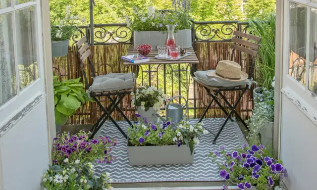 Gemütliche Balkon Ideen: Ein kleiner Tisch mit Getränken und Kirschen, zwei Stühle mit Kissen, Blumen und eine Gießkanne laden zum Verweilen ein.