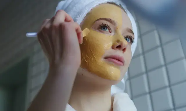 Junge Frau mit Handtuch auf dem Kopf trägt eine Kurkuma-Maske auf. Die Kurkuma Wirkung auf die Haut ist bekannt für ihre entzündungshemmenden Eigenschaften.