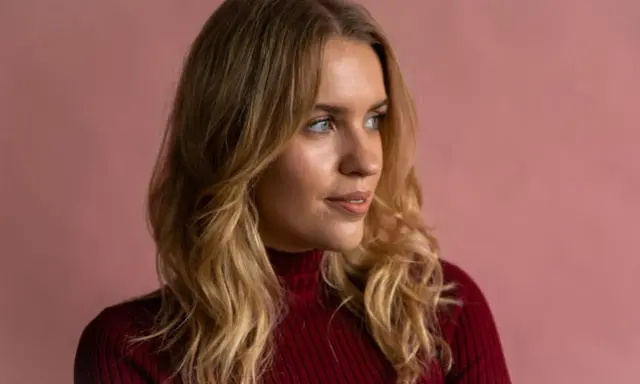 Blonde Frau mit einem **kitty cut**-Haarschnitt, trägt einen roten Rollkragenpullover vor rosafarbenem Hintergrund.