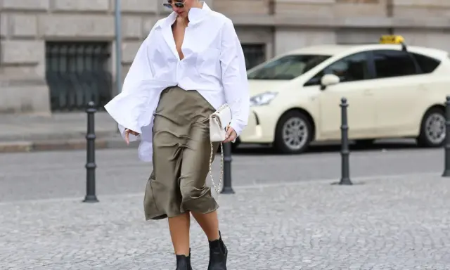 Eine Frau stylt eine weiße Oversize Bluse mit einem Satinrock und schwarzen Stiefeln. Sie trägt eine weiße Tasche.