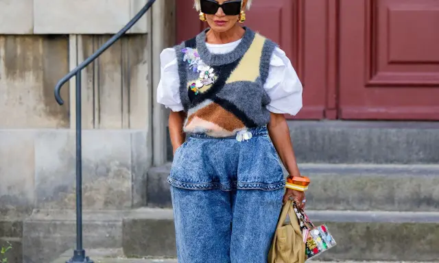 Modische Frau mit weißem Haar, Sonnenbrille und auffälligem Pullunder, der lässig zu Jeans kombiniert wird.