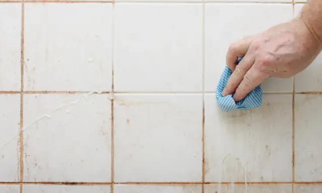 Hand reinigt schmutzige weiße Wandfliesen mit einem blauen Tuch.