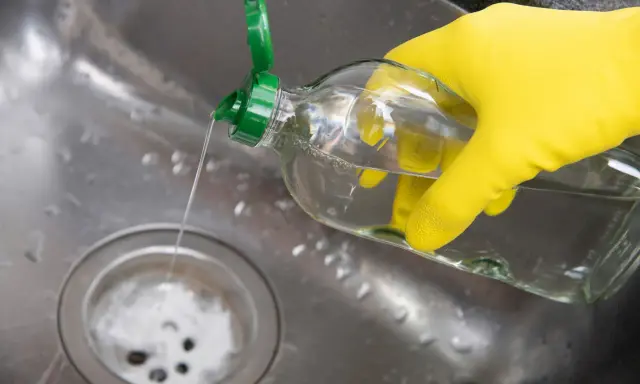Gelbe Handschuhe halten eine Flasche mit klarem Flüssigkeit, die in ein Spülbecken gegossen wird. Ein Hausmittel Rohrreiniger, der Abfluss wird gereinigt.