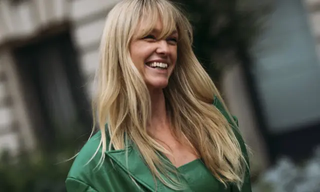Lachende Frau mit **leichter Pony, lange Haare** und einem grünen Blazer.