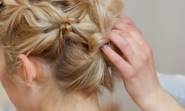 Eine Hand stylt blonde Haare zu verspielten hochsteckfrisuren mittellang. Kleine Zöpfe und gedrehte Strähnen ergeben einen lässigen Look.