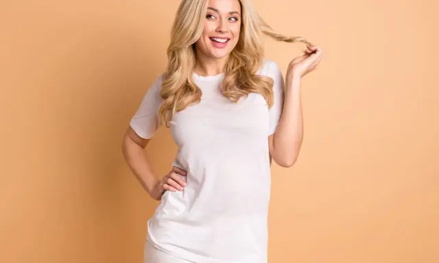 Eine lächelnde Vanilla Girl mit blonden Locken spielt mit einer Haarsträhne. Sie trägt ein weißes T-Shirt und weiße Hose.
