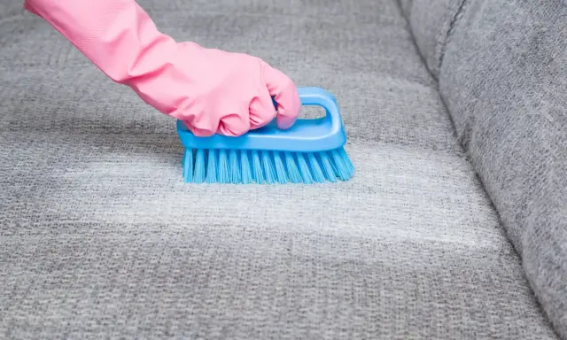 Hand in pink glove uses blue brush to scrub grey fabric, showing how to remove blutflecken.