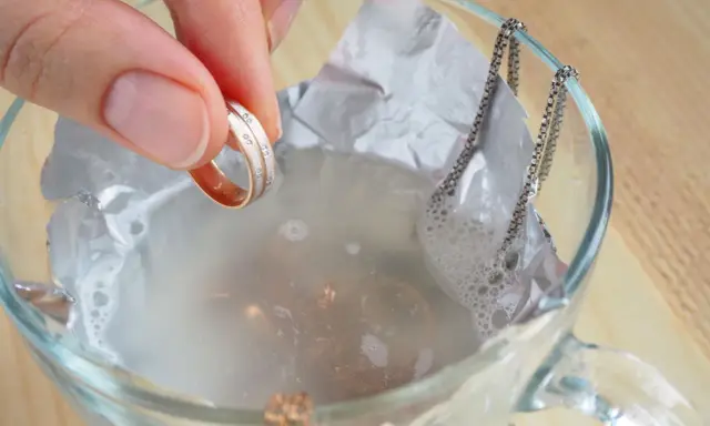 Schmuckreinigung mit Hausmitteln: Eine Hand hält einen Ring, während eine Silberkette in einer Schüssel mit Wasser und Alufolie liegt.