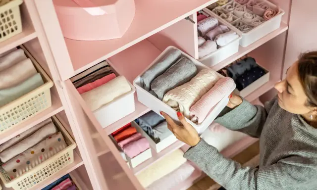 Frau sortiert Kleidung in einem rosa Kleiderschrank. Sie zeigt, wie man den Kleiderschrank organisiert, mit gefalteten Pullovern in weißen Boxen.