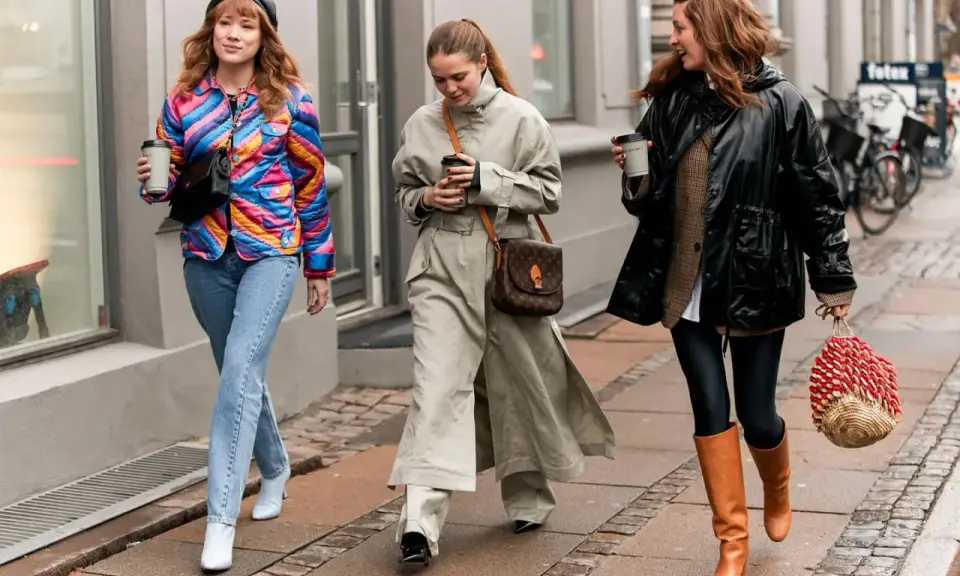 Drei Frauen spazieren mit Kaffee. Die mittlere trägt weite Hosen zu Pumps, die rechte Leggings zu Stiefeln. Die linke zeigt, welche Hose zu Boots Damen passt: Jeans zu weißen Stiefeletten.