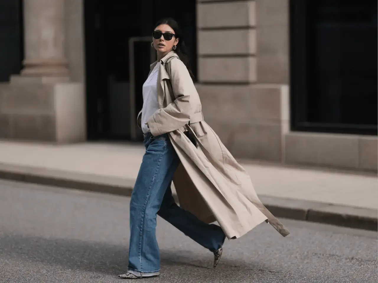 Eine Frau im Trenchcoat und Jeans, Sonnenbrille, Schmuck. Ihr Stil ist klassisch und clean girl.