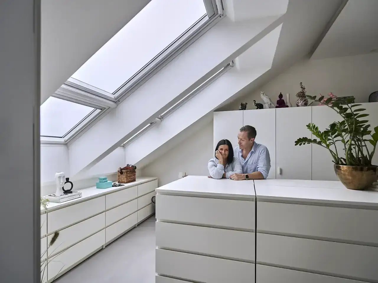 Ein Paar in einem hellen Ankleidezimmer mit Dachfenstern. Viele weiße Schubladen bieten tolle Ankleidezimmer Ideen für Stauraum.