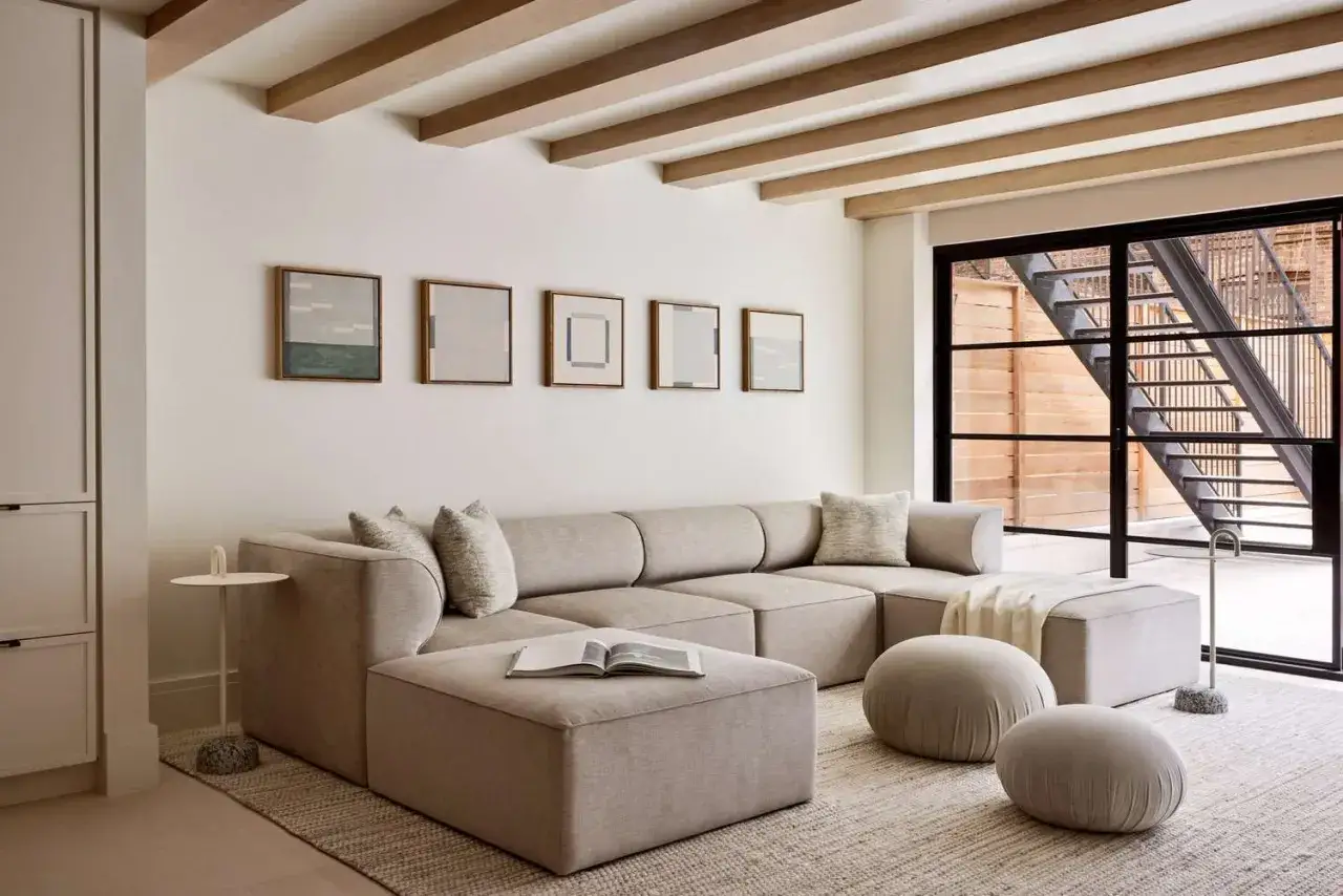 Einladendes, modernes Wohnzimmer mit beigem Ecksofa, Poufs und Kunst an der Wand. Holzbalken an der Decke und ein großes Fenster mit Außentreppe.