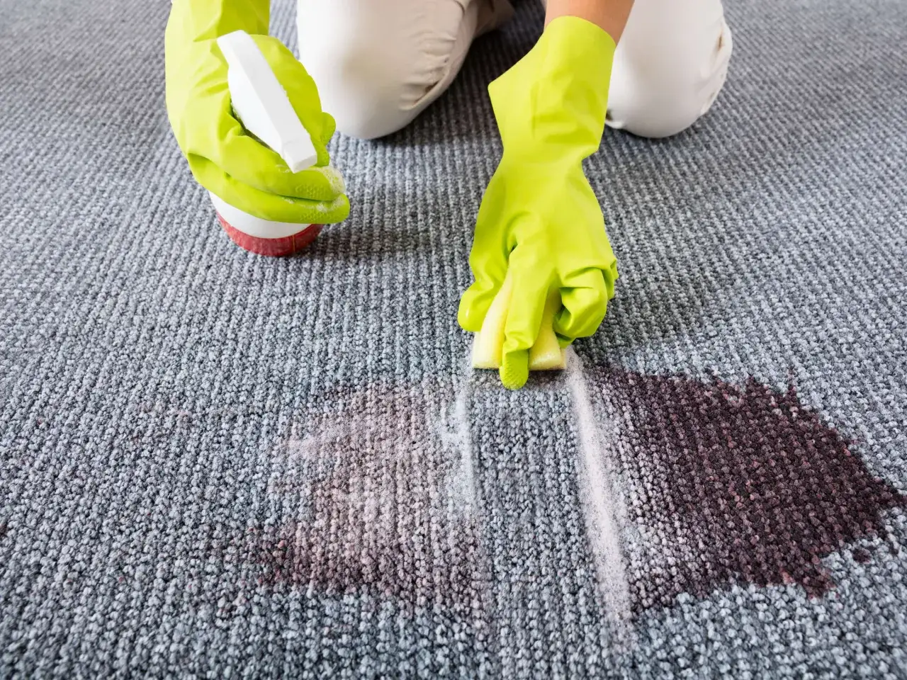 H&auml;nde in gr&uuml;nen Handschuhen spr&uuml;hen Reiniger auf einen Fleck und reiben ihn mit einem Schwamm ein, um den Teppichboden zu reinigen.
