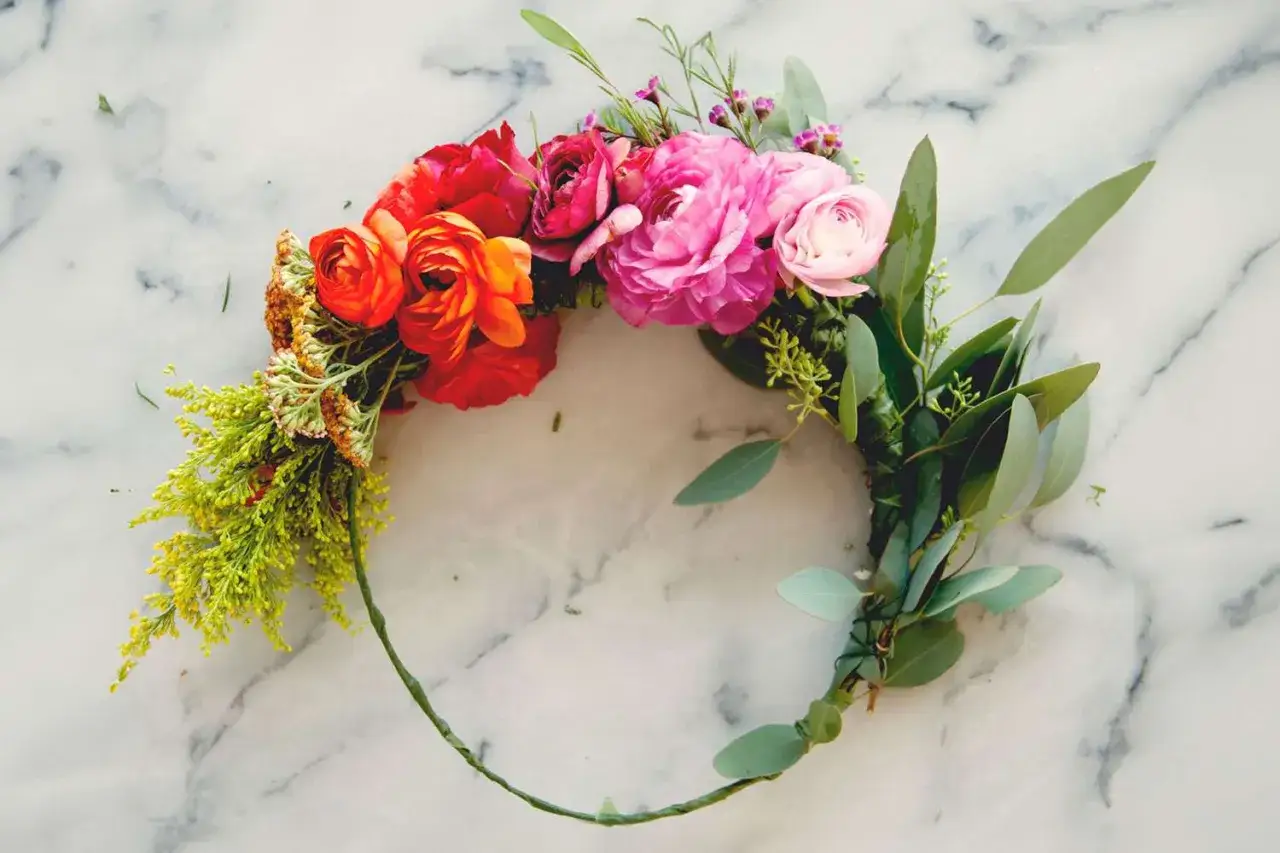 Ein bunter Blumenkranz wird gebunden, mit roten, orangen und pinken Blüten sowie grünen Blättern auf Marmor.