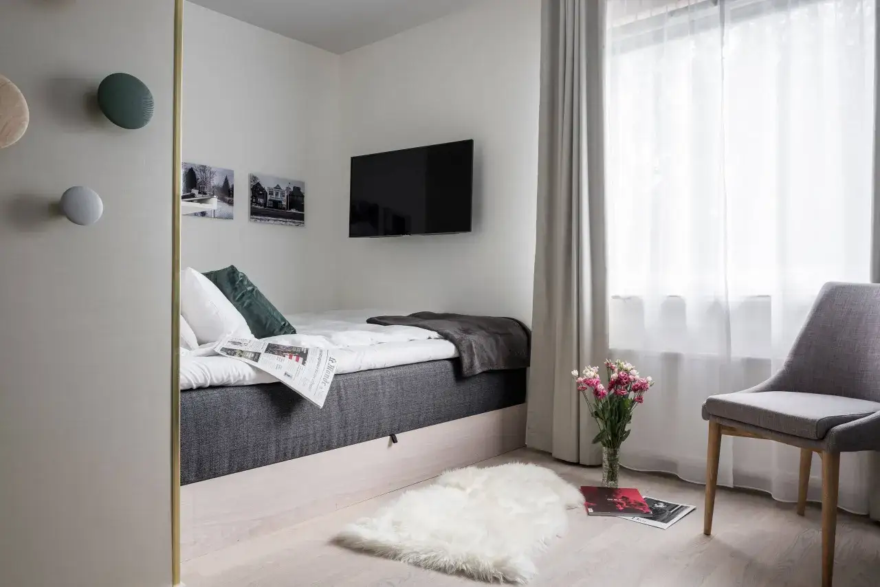 Gem&uuml;tliches Schlafzimmer mit einem modernen Fernseher an der Wand, einem gemachtem Bett mit Kissen und Decke, sowie einem Fenster mit Vorh&auml;ngen.