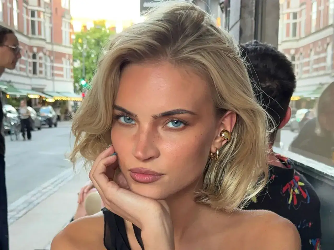 Blonde Frau mit Scandi Bob und blauen Augen, die nachdenklich in die Kamera blickt. Goldener Schmuck.
