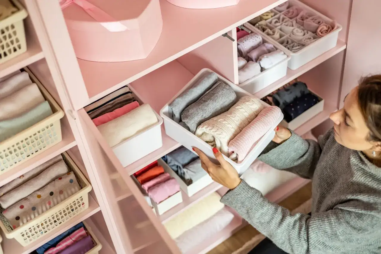 Frau sortiert Kleidung in einem rosa Kleiderschrank. Sie zeigt, wie man den Kleiderschrank organisiert, mit gefalteten Pullovern in Beh&auml;ltern.