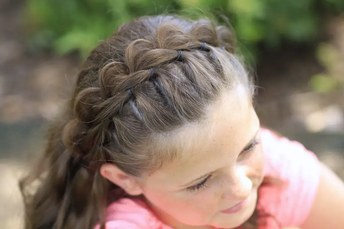 Ein Mädchen mit braunen Haaren trägt eine geflochtene Frisur mit Haarband. Kleine Zöpfe werden mit Haargummis gehalten und bilden einen Kranz.