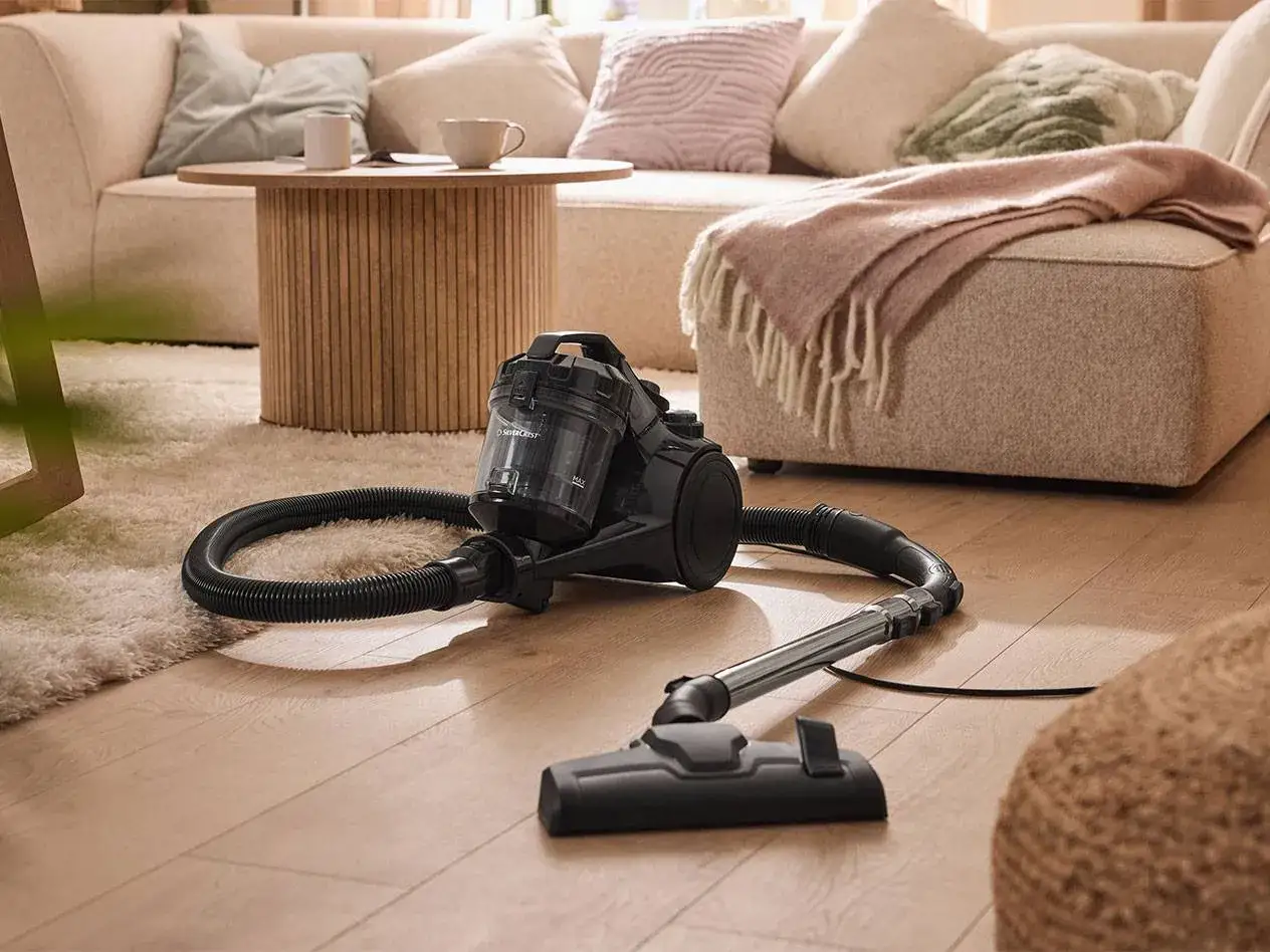 Staubsauger bereit für die couch reinigen. Ein moderner Staubsauger mit Schlauch und Bürste liegt auf dem Holzboden vor einer gemütlichen Couch.