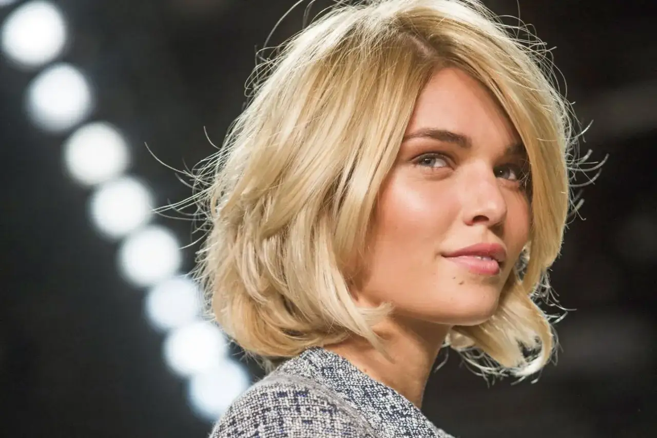 Blonde Frau mit einem l&auml;ssigen Scandi Bob und einem Tweed-Jackett, im Hintergrund unscharfe Lichter.