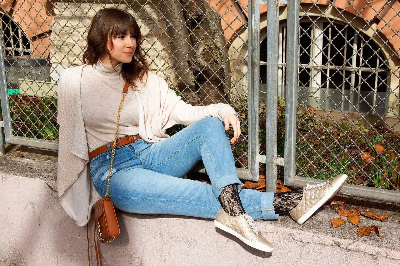 Frau sitzt lässig in hellblauen Mom Jeans, kombiniert mit einem cremefarbenen Rollkragenpullover und einer Umhängetasche. Goldene Sneaker und Netzstrümpfe runden den Look ab.